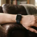 A close-up of a hand wearing a sleek, minimalist smartwatch, resting casually on a rich, dark leather armchair armrest